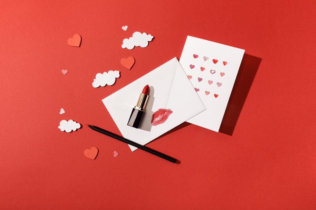 bovenaanzicht van papieren wolken en harten, wenskaart nabij enveloppe met lipprint, lippenstift en potlood op rode achtergrond - Foto, afbeelding