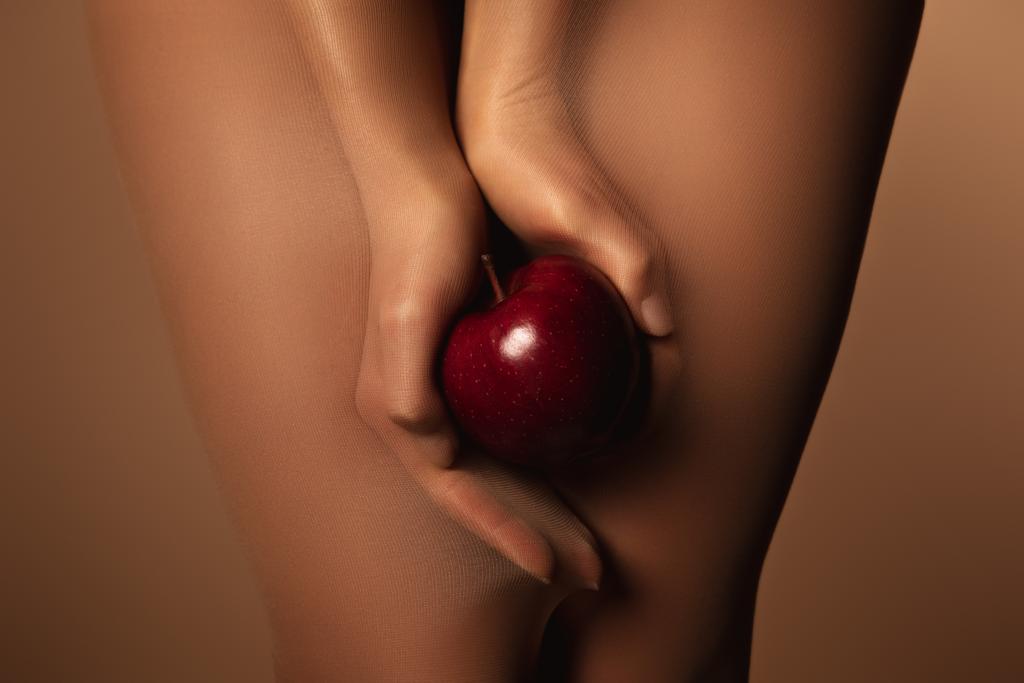 abgeschnittene Ansicht der Frau in Nylonstrumpfhosen mit reifem roten Apfel isoliert auf braun - Foto, Bild