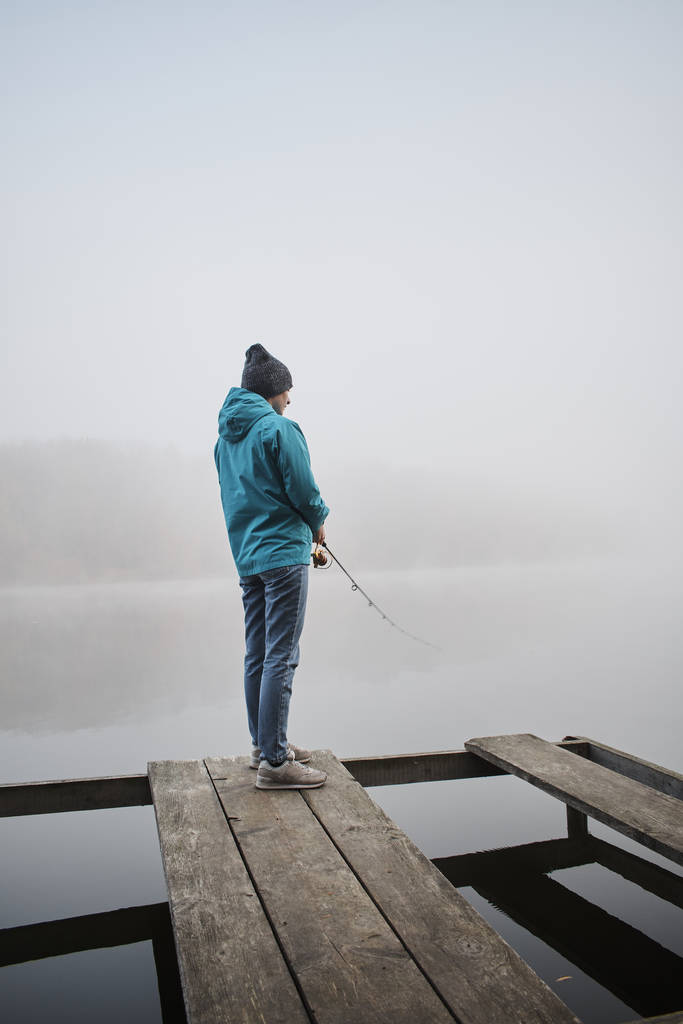 Rückansicht einer jungen Frau, die am nebligen Morgen auf einem Holzsteg steht und mit der Angel fischt - Foto, Bild