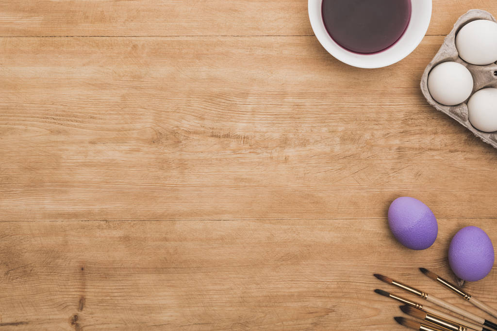 vista superior de acuarela pintura púrpura en tazón cerca de huevos de pollo y pinceles en mesa de madera
 - Foto, Imagen
