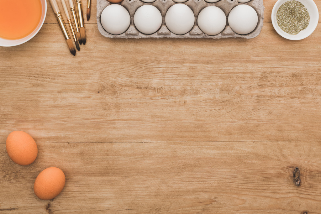 vista dall'alto di vernice arancione ad acquerello e brillantini dorati in ciotole vicino alle uova di Pasqua e pennelli sul tavolo di legno
 - Foto, immagini