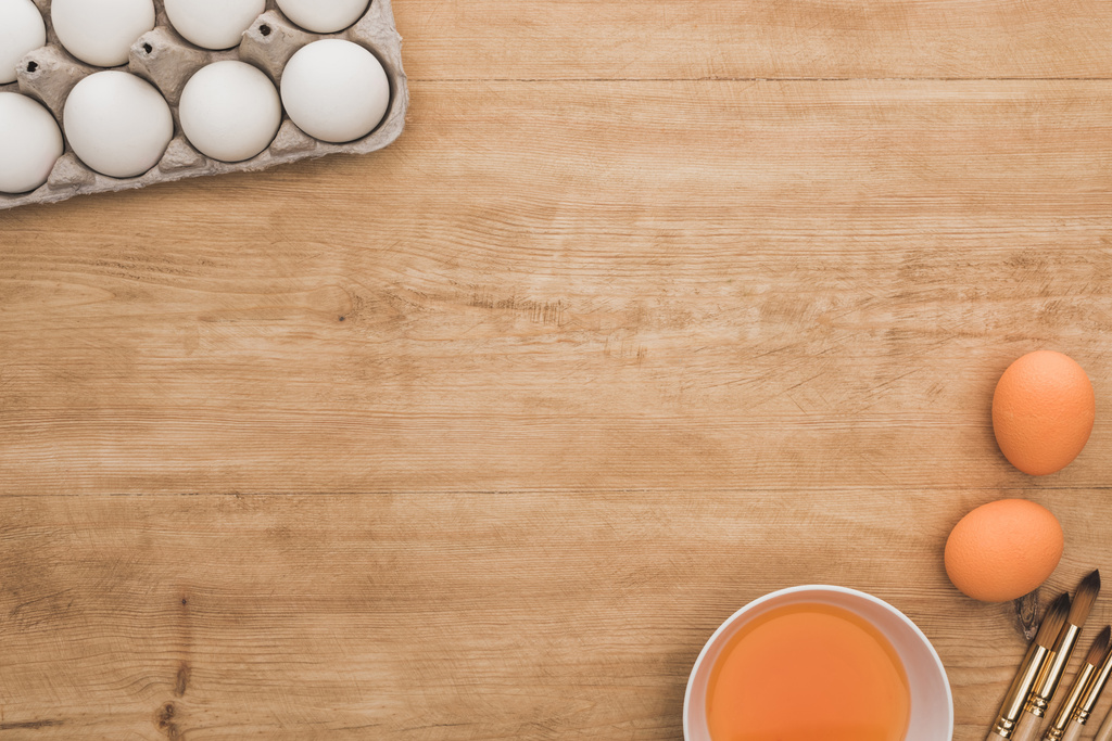 鶏の卵の近くのボウルにオレンジの水彩画のトップビューと木製のテーブルの上の絵筆 - 写真・画像
