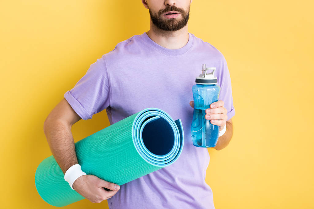cropped view of bearded man holding sports bottle and fitness mat on yellow - 写真・画像