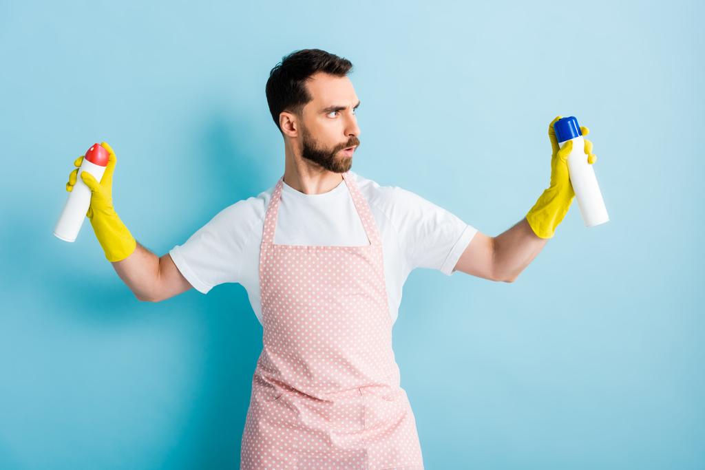 серйозний бородатий чоловік тримає пляшки розпилення на синьому
  - Фото, зображення