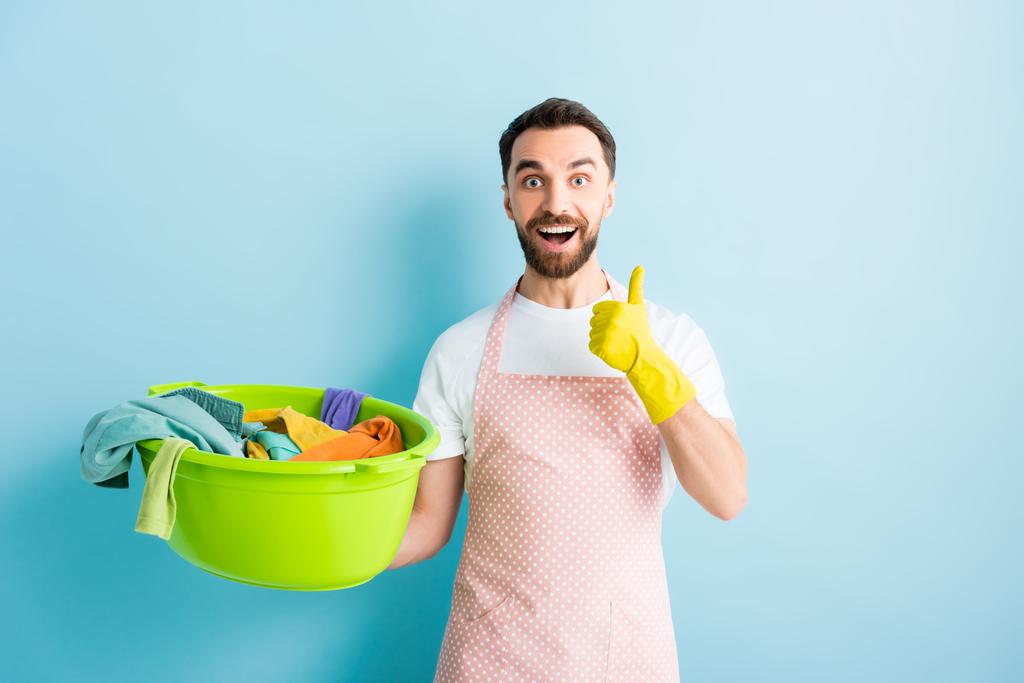 happy bearded man holding plastic wash bowl with dirty laundry and showing thumb up on blue  - Photo, Image