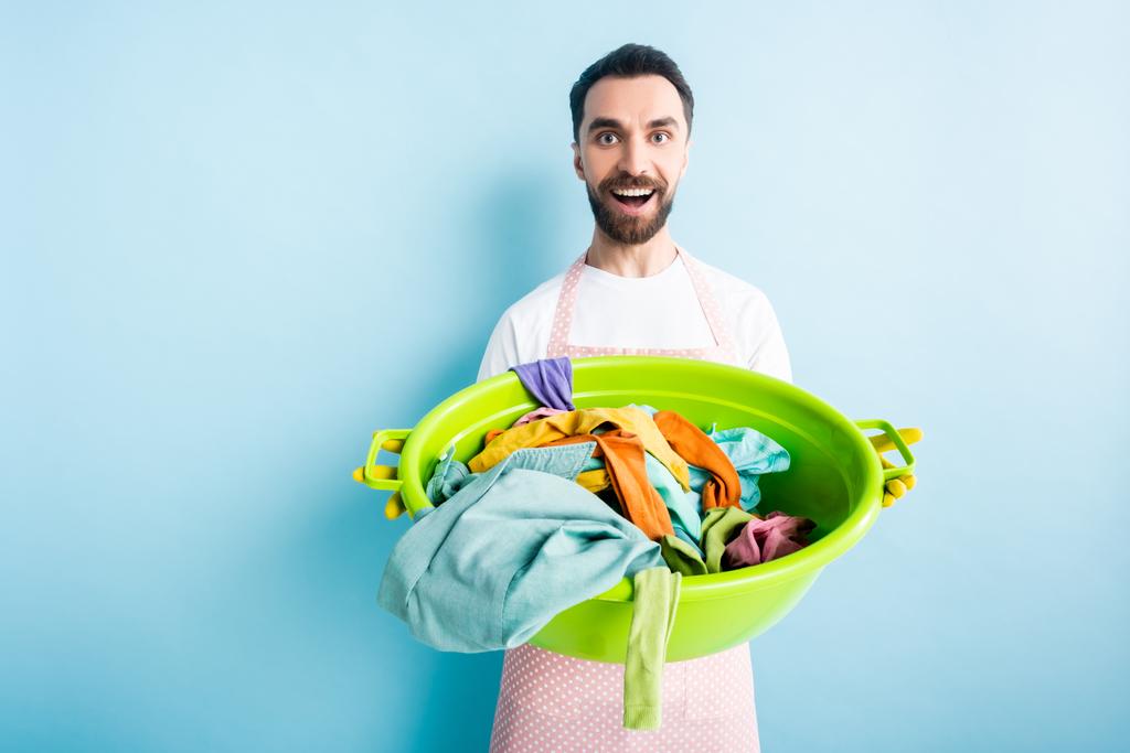 cheerful bearded man holding dirty laundry in plastic wash bowl on blue  - Photo, Image