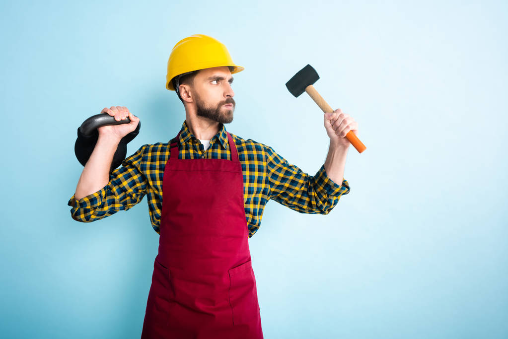 naštvaný vousatý dělník v bezpečnostní přilbě drží kladivo a činka na modré  - Fotografie, Obrázek