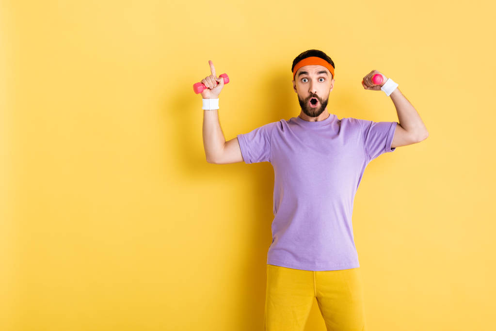 surprised bearded man pointing with finger while holding small dumbbells on yellow - Photo, Image