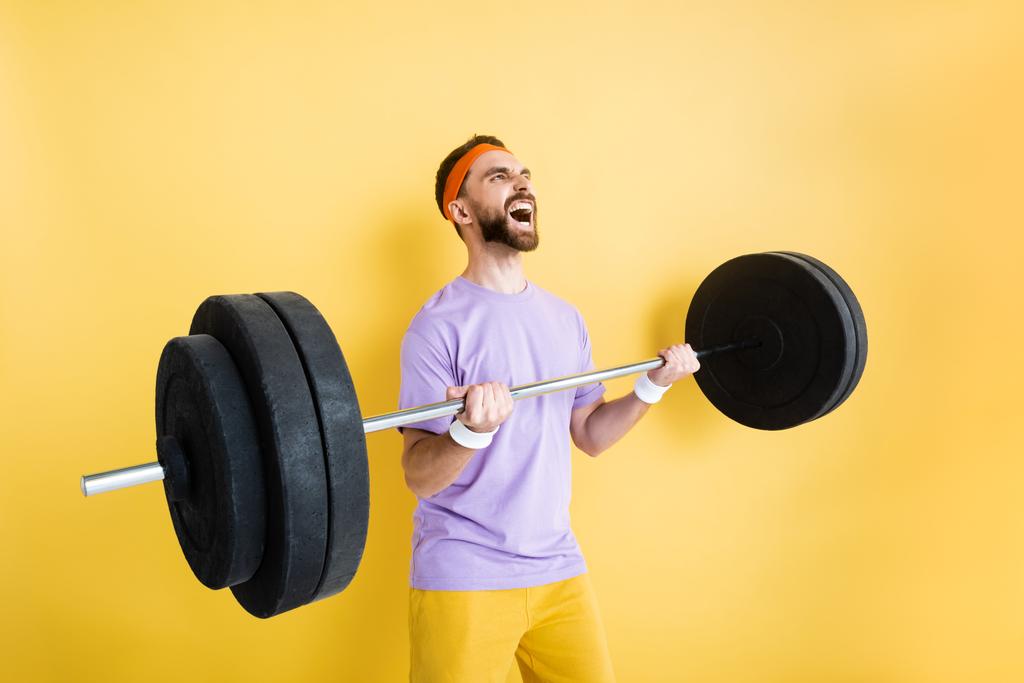 emotional bearded sportsman working out with barbell on yellow - Photo, Image