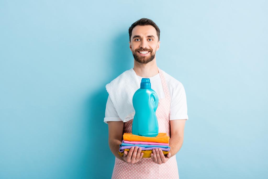 šťastný vousatý muž drží čisté oblečení a prací prášek na modré  - Fotografie, Obrázek