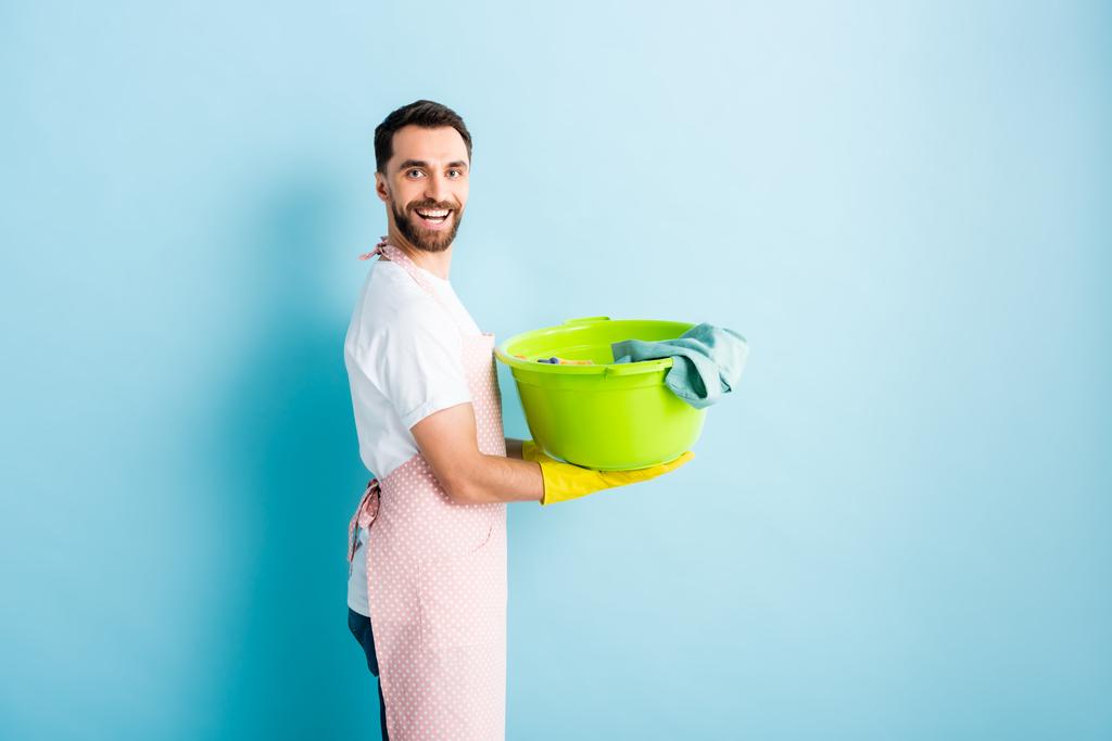 позитивний бородатий чоловік в фартусі тримає брудну білизну на синьому
  - Фото, зображення