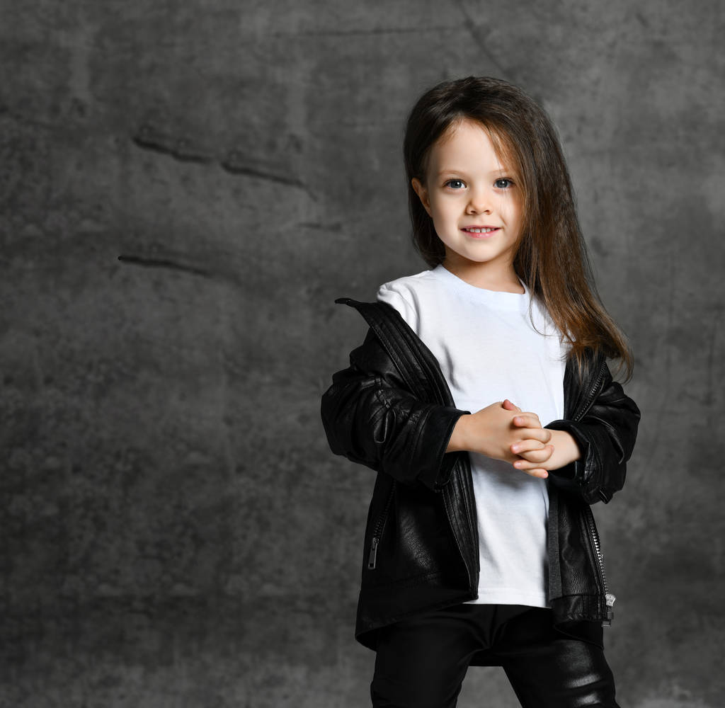 Pieni hymyilevä söpö tyttö mustavalkoinen rock-tähti tyyli rento vaatteet seisoo ja katselee kameraa yli harmaa betoni tausta
 - Valokuva, kuva