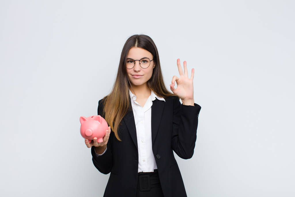 young pretty woman feeling happy, relaxed and satisfied, showing approval with okay gesture, smiling with a piggy bank - Photo, Image
