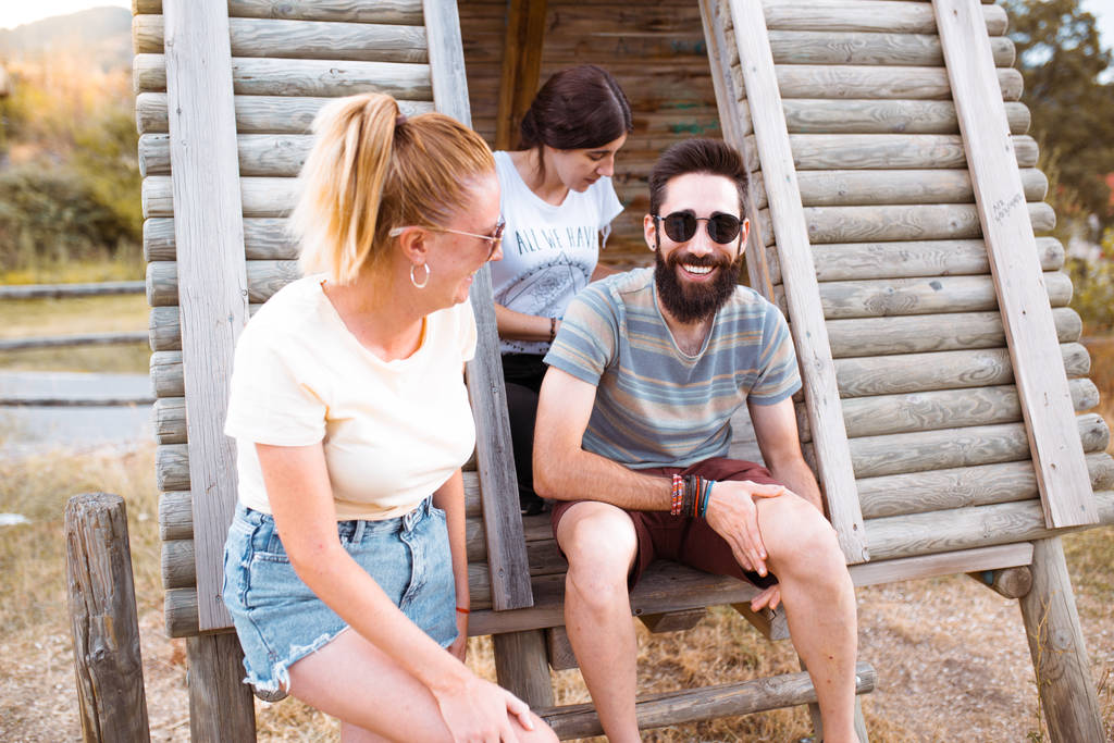 Turyści się relaksują. Grupa trzech przyjaciół siedzi na drewnianych schodach obozowych ciesząc się i śmiejąc na wsi. Przygody obozowe i koncepcja podróży. - Zdjęcie, obraz
