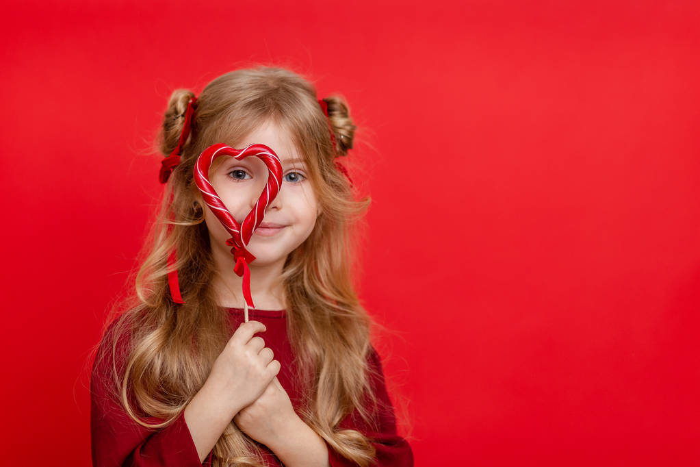 Retrato de uma menina sonhadora alegre com um doce em forma de coração em suas mãos isolado em um fundo vermelho
 - Foto, Imagem