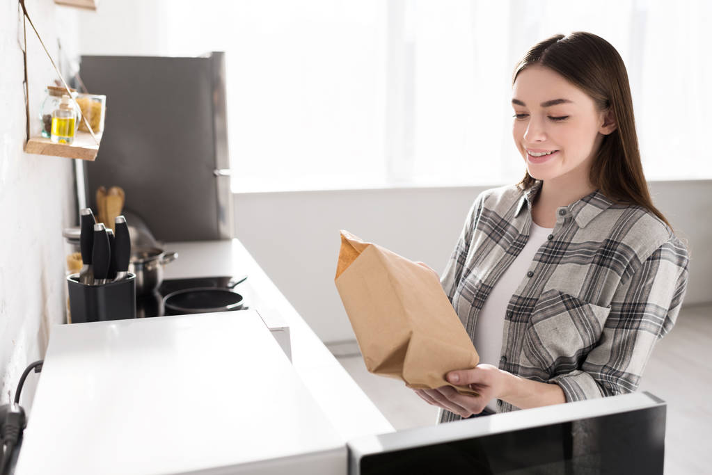 usmívající se žena drží papírový sáček s popcornem v blízkosti mikrovlnky v kuchyni  - Fotografie, Obrázek