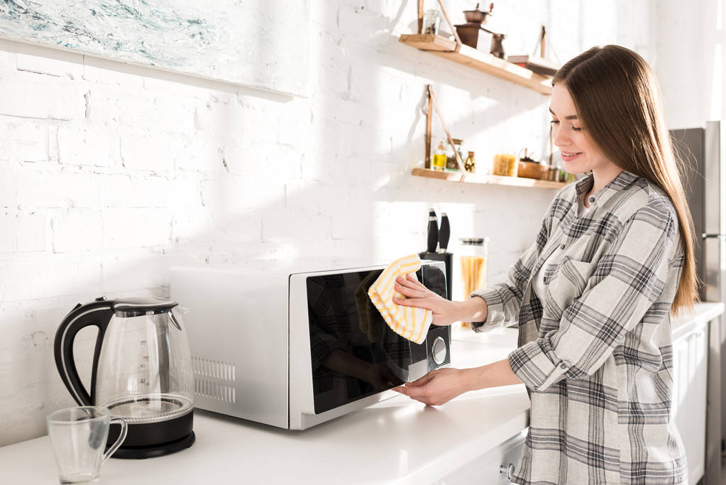 mulher sorridente e atraente limpeza de microondas com pano na cozinha
  - Foto, Imagem