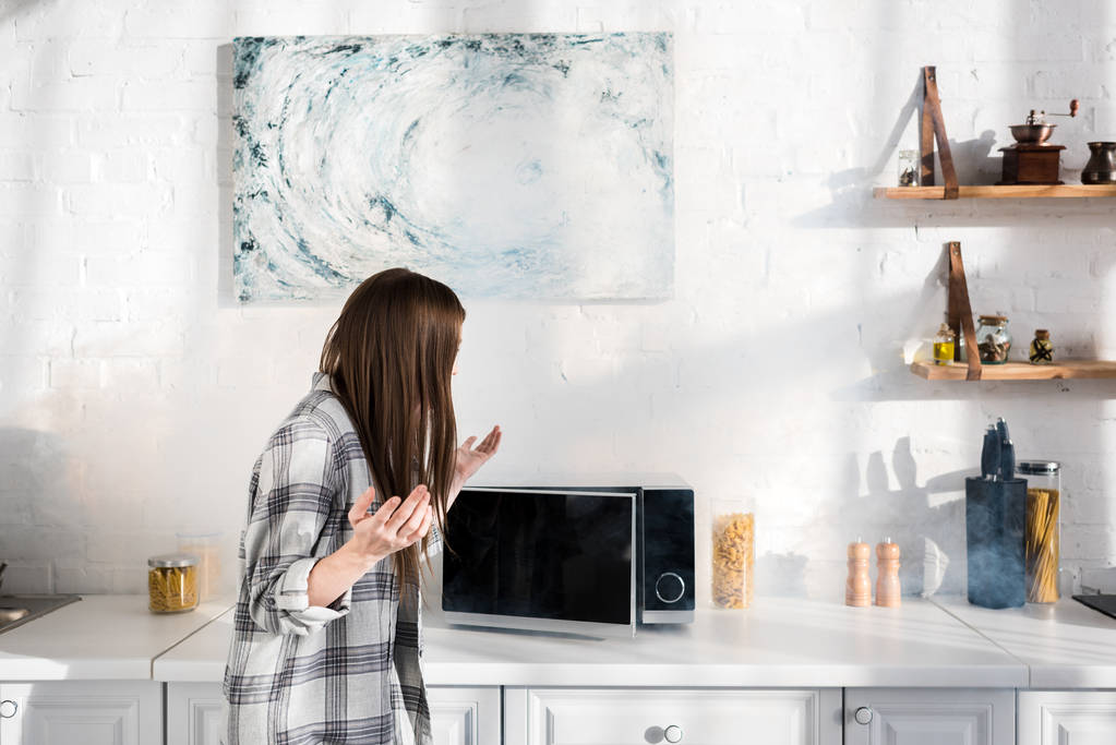 Schockierte Frau blickt auf kaputte Mikrowelle in Küche  - Foto, Bild