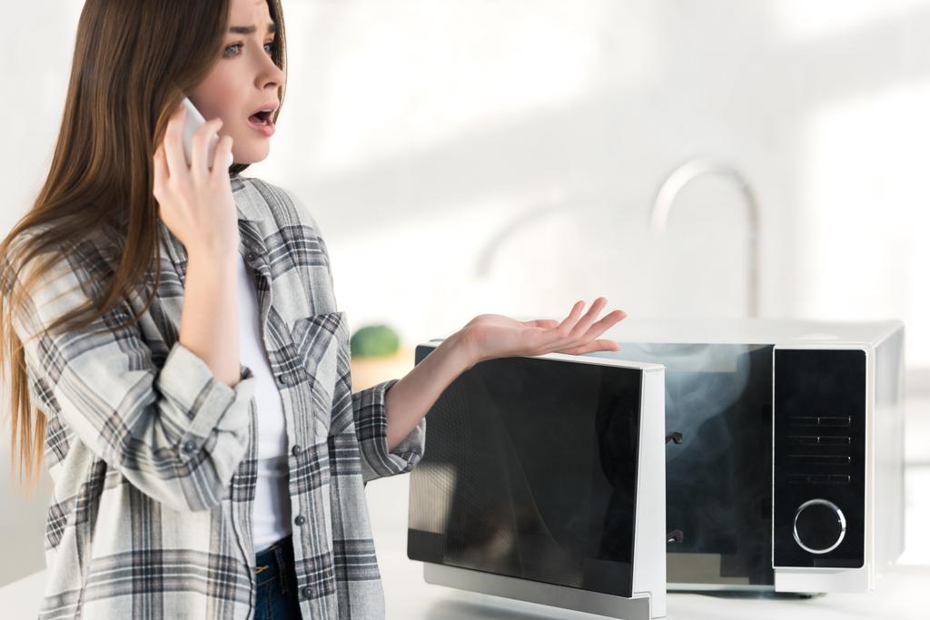 donna scioccata che parla su smartphone vicino al microonde rotto in cucina
  - Foto, immagini