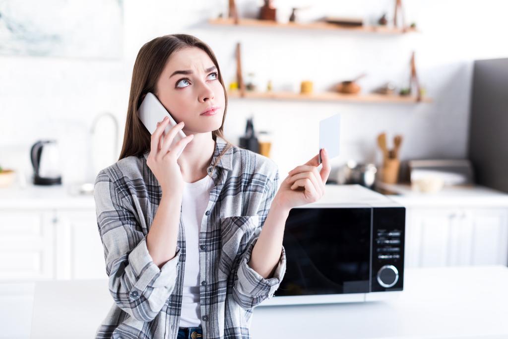 mujer enojada hablando en el teléfono inteligente cerca de microondas y celebración de la tarjeta de visita en la cocina
  - Foto, Imagen