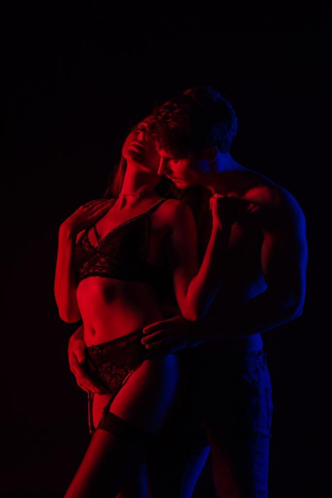man touching undressed sexy young woman in lingerie in red light isolated on black - Photo, Image
