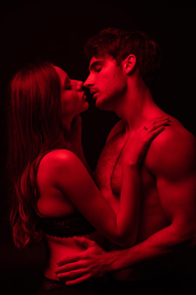 вид сбоку страстной раздетой сексуальной молодой пары целующейся в красном свете изолированной на черном
 - Фото, изображение