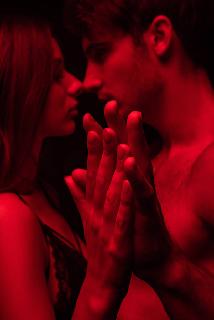 вид сбоку страстной раздетой сексуальной молодой пары целующейся и держащейся за руки в красном свете изолированной на черном
 - Фото, изображение