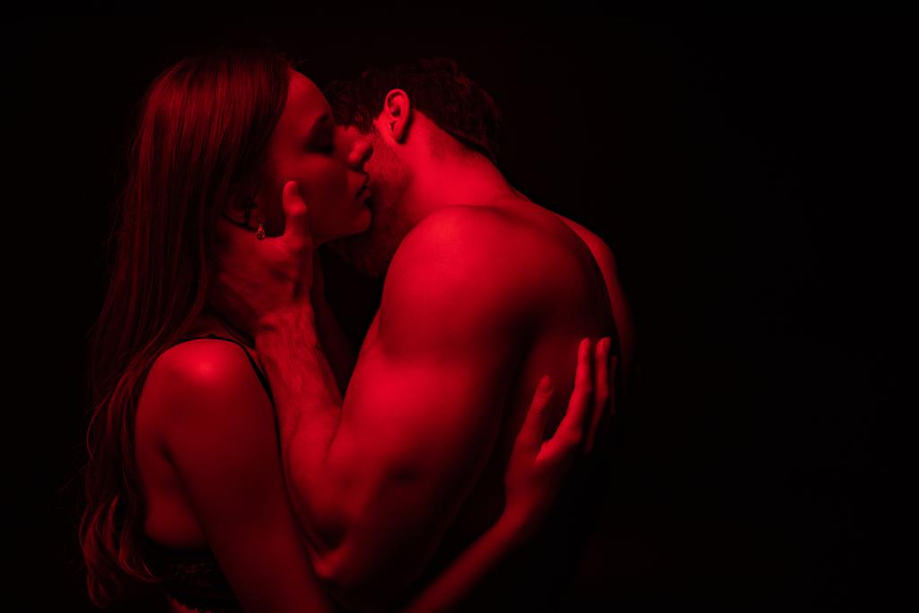 Kırmızı ışıkta öpüşen, tutkulu, çıplak ve seksi genç çiftin yan görünüşü siyahta izole. - Fotoğraf, Görsel