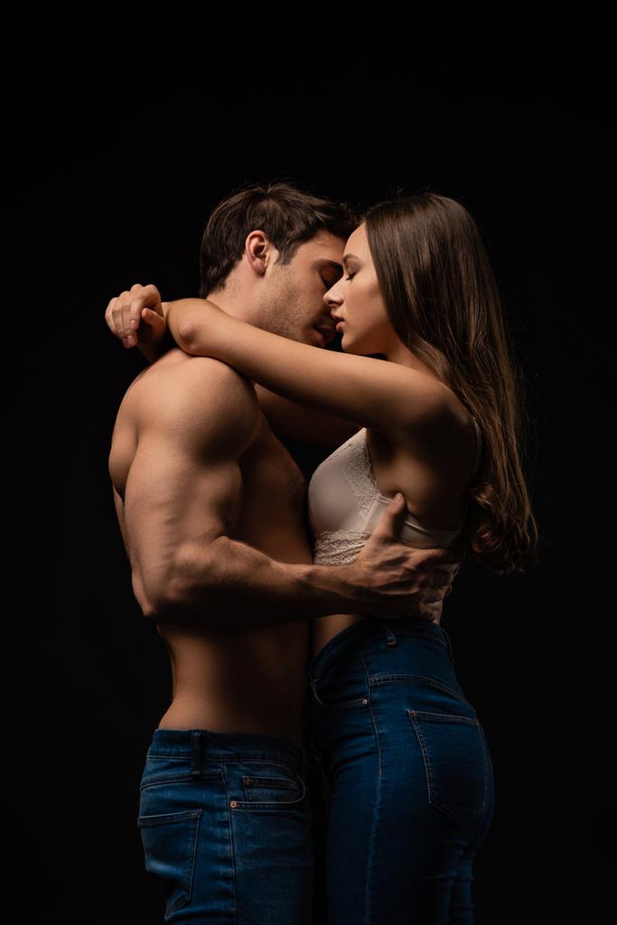 vista lateral de vestido sexy jovem casal em jeans abraçando e beijando isolado no preto
 - Foto, Imagem
