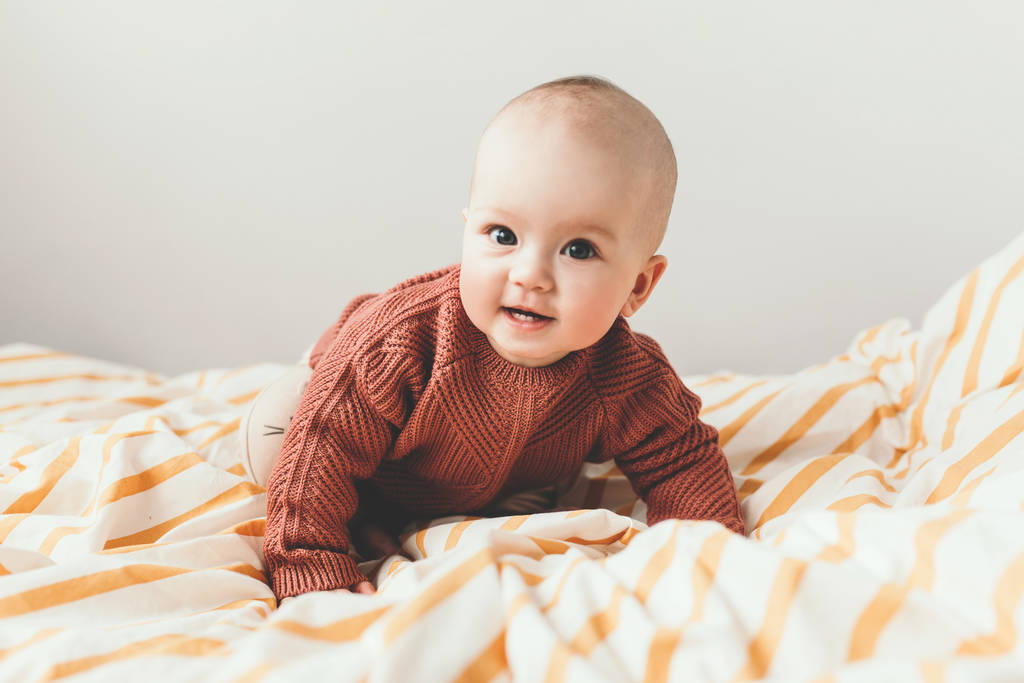 Krásná holčička na posteli v útulném hnědém svetru se usmívá. Pojem mateřství a dětství - Fotografie, Obrázek