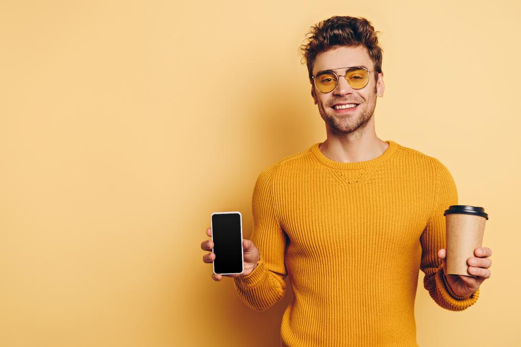 knappe man met smartphone met leeg scherm terwijl het houden van koffie te gaan op gele achtergrond - Foto, afbeelding