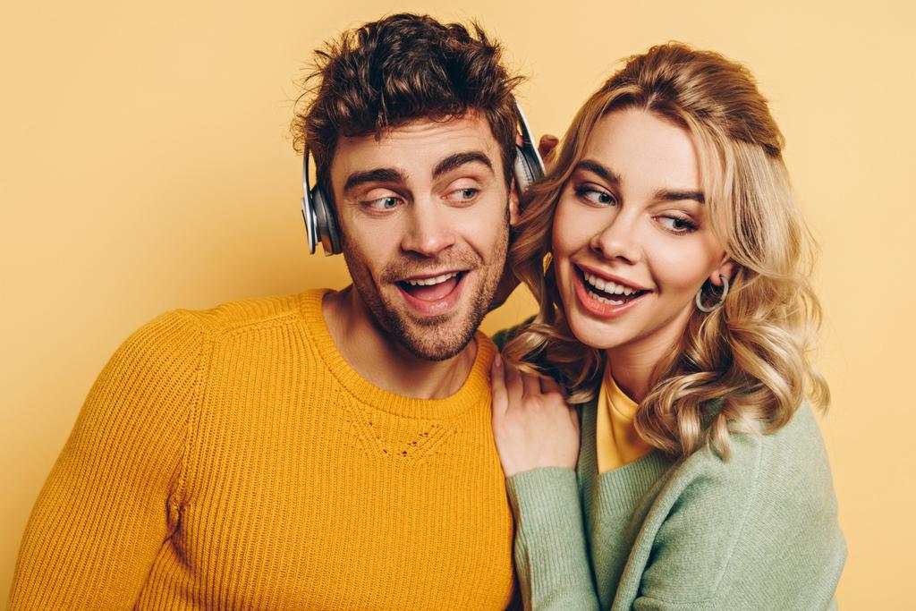 Mutlu kız, sarı arka planda kablosuz kulaklıkla müzik dinlerken erkek arkadaşını kucaklıyor. - Fotoğraf, Görsel