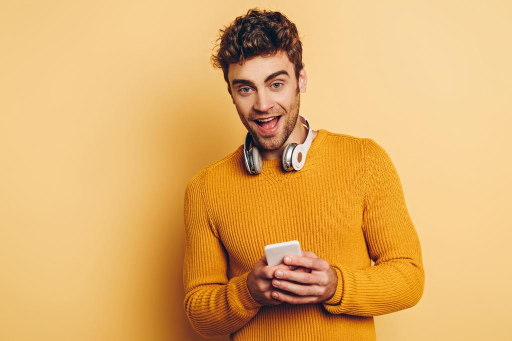 όμορφος άνδρας με ασύρματα ακουστικά στο λαιμό χρησιμοποιώντας smartphone και χαμογελώντας στην κάμερα σε κίτρινο φόντο - Φωτογραφία, εικόνα