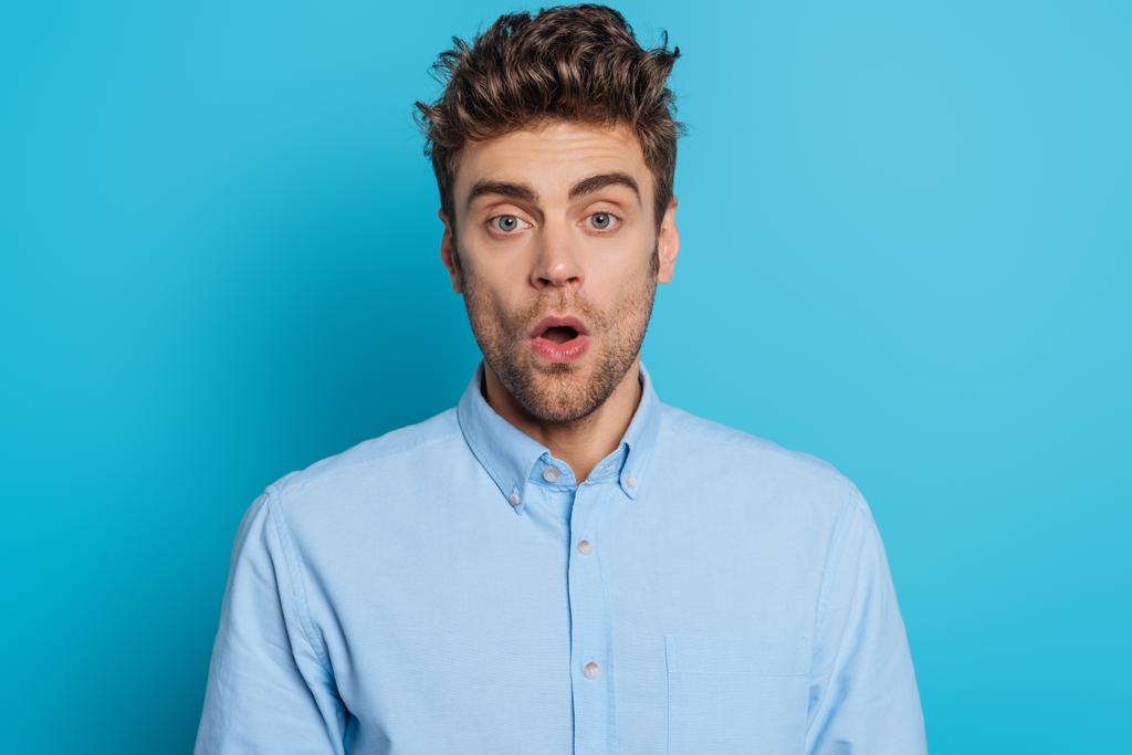 Schockierter junger Mann blickt mit offenem Mund auf blauem Hintergrund in die Kamera - Foto, Bild