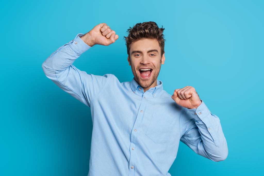 возбужденный молодой человек показывает жест победителя, улыбаясь в камеру на синем фоне
 - Фото, изображение