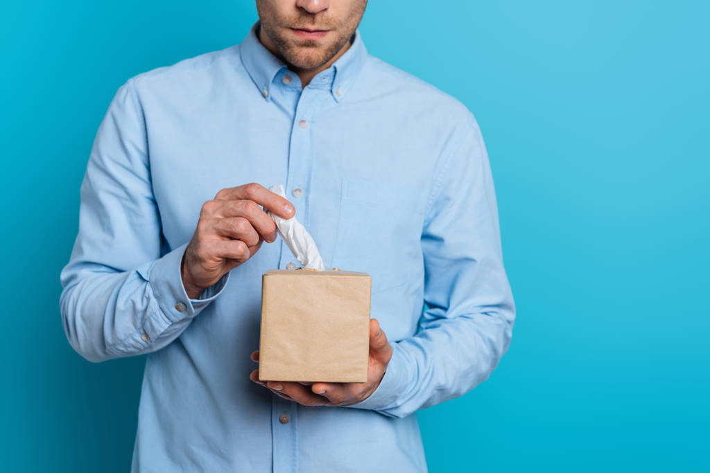 обрезанный вид расстроенного молодого человека, берущего бумажную салфетку из упаковки на синем фоне
 - Фото, изображение