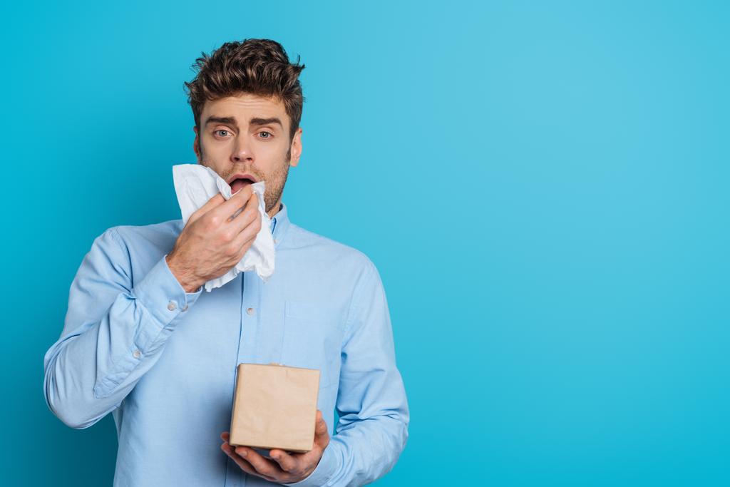 sick man sneezing in paper napkin while looking at camera on blue background - Photo, Image