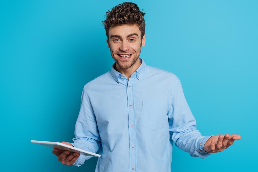 веселый молодой человек, стоящий с открытой рукой, держа в руках цифровой планшет и улыбаясь в камеру на синем фоне
 - Фото, изображение