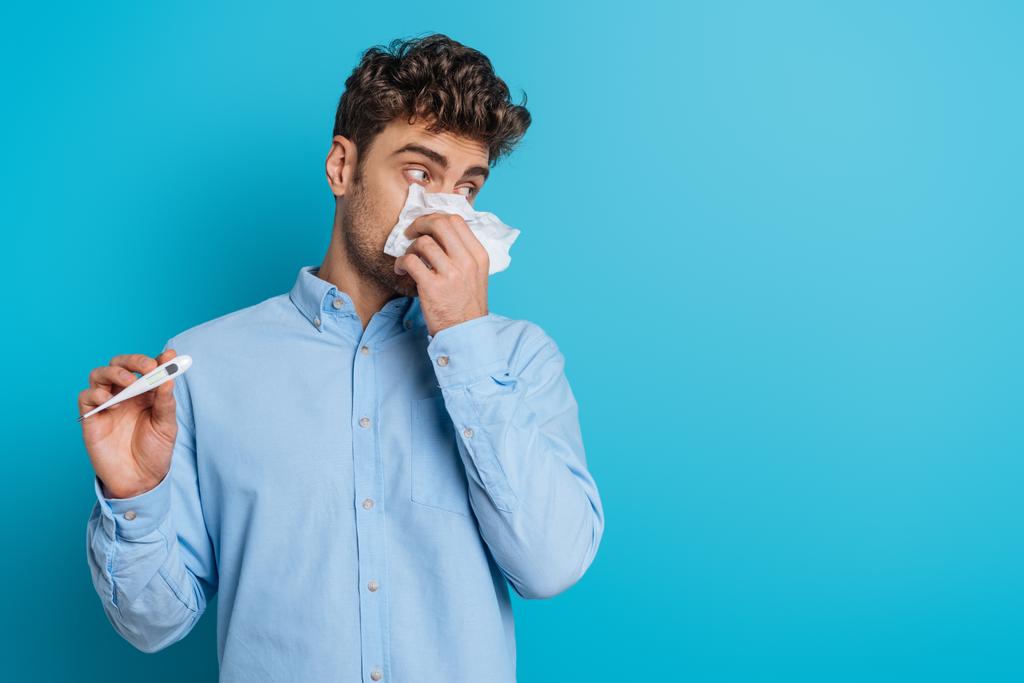 chory młody człowiek wycierający nos papierową serwetką i pokazujący termometr na niebieskim tle - Zdjęcie, obraz