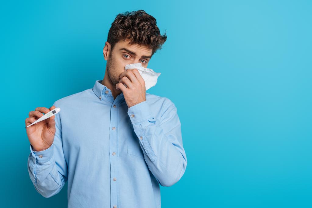άρρωστος νεαρός άνδρας σκουπίζοντας τη μύτη με χαρτοπετσέτα και δείχνοντας θερμόμετρο σε μπλε φόντο - Φωτογραφία, εικόνα