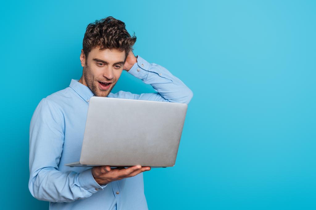 έκπληκτος νεαρός άνδρας αγγίζοντας το κεφάλι, ενώ κοιτάζοντας το φορητό υπολογιστή σε μπλε φόντο - Φωτογραφία, εικόνα