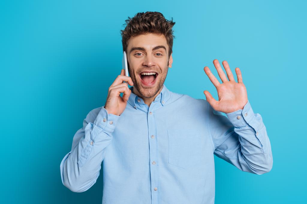 happy young man waving hand while talking on smartphone on blue background - Photo, Image