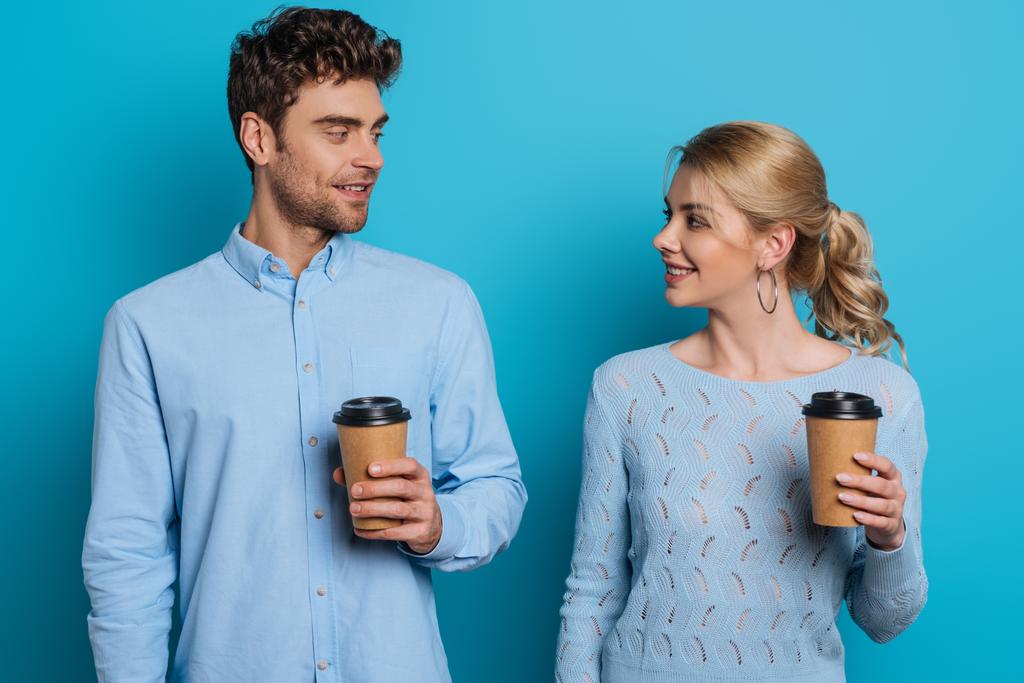 smiling man and woman holding disposable cups while looking at each other on blue background - Photo, Image