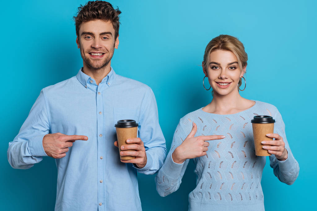 улыбающиеся мужчина и женщина указывая пальцами на одноразовые чашки, глядя на камеру на синем фоне
 - Фото, изображение