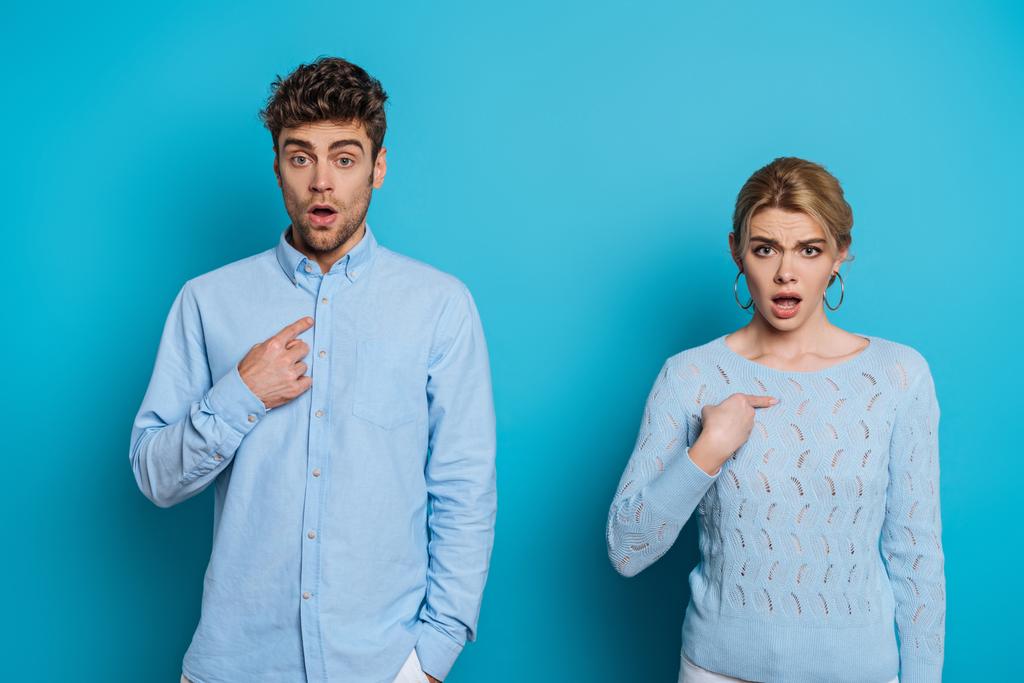 šokovaný muž a žena ukazuje prsty na sebe při pohledu na kameru na modrém pozadí - Fotografie, Obrázek