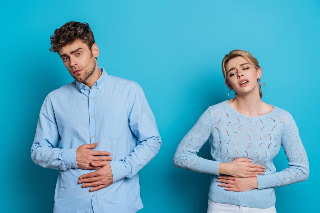 perturbado homem e mulher tocando estômagos enquanto sofre de dor abdominal no fundo azul
 - Foto, Imagem