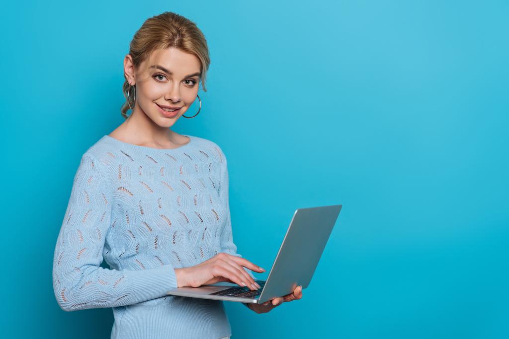 cheerful girl smiling at camera while using laptop on blue background - Photo, Image