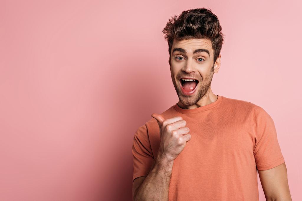 excited man pointing with thumb while looking at camera on pink background - Photo, Image