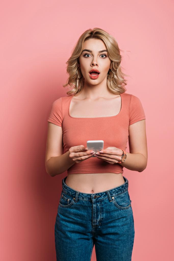 шокированная девушка смотрит в камеру во время чата на смартфоне на розовом фоне
 - Фото, изображение