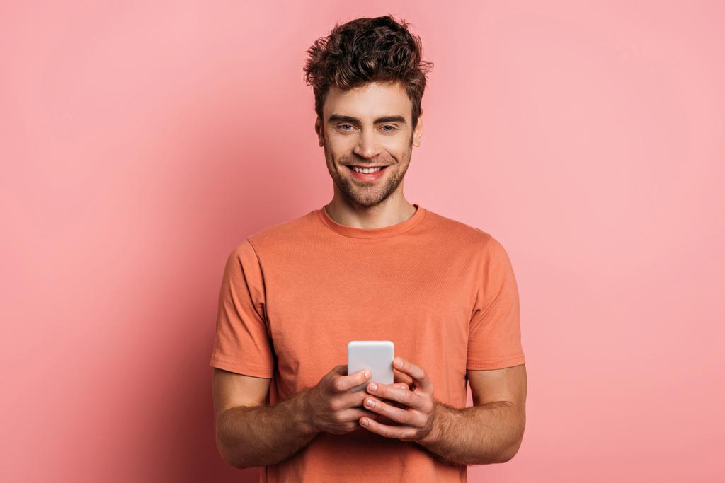 positive young man smiling at camera while chatting on smartphone on pink background - 写真・画像